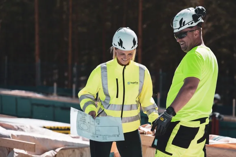 Smilende prosjektleder og taktekker på arbeidsplassen, med arbeidsklær og hjelm
